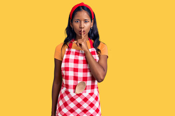 Young indian girl wearing professional baker apron asking to be quiet with finger on lips. silence and secret concept.