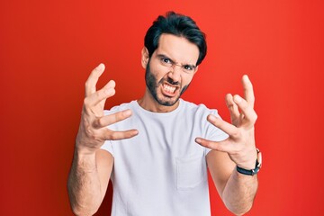 Fototapeta premium Young hispanic man wearing casual white tshirt shouting frustrated with rage, hands trying to strangle, yelling mad