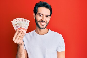 Young hispanic man holding 10 colombian pesos banknotes looking positive and happy standing and smiling with a confident smile showing teeth