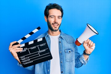 Young hispanic man holding video film clapboard and megaphone winking looking at the camera with...