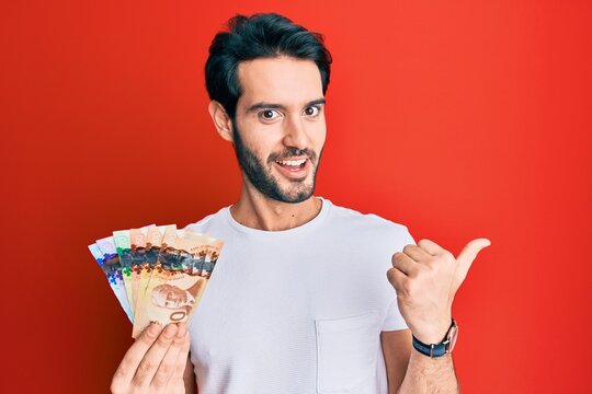 Young hispanic man holding canadian dollars pointing thumb up to the side smiling happy with open mouth