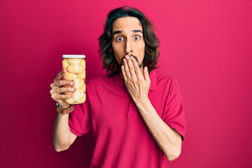 Young hispanic man holding bottle with boiled potatoes covering mouth with hand, shocked and afraid...