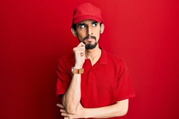 Young hispanic man wearing delivery uniform and cap serious face thinking about question with hand on chin, thoughtful about confusing idea