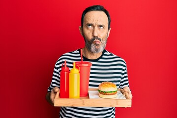 Middle age hispanic man eating a tasty classic burger and soda making fish face with mouth and squinting eyes, crazy and comical.