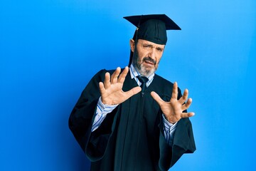 Middle age hispanic man wearing graduation cap and ceremony robe afraid and terrified with fear expression stop gesture with hands, shouting in shock. panic concept.