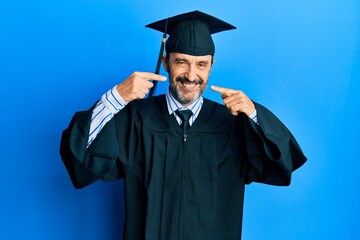 Middle age hispanic man wearing graduation cap and ceremony robe smiling cheerful showing and pointing with fingers teeth and mouth. dental health concept.