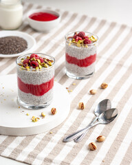 Two glass jar of chia pudding with raspberry, pistachio and red jam on a white table with striped tablecloth.