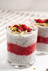 Transparent glass jar of chia pudding with raspberry, pistachio and red jam on a white table with striped tablecloth.