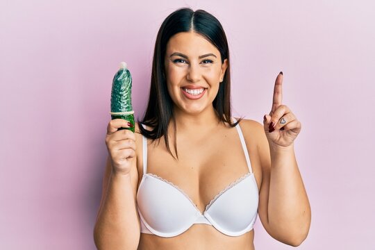 Beautiful brunette woman holding condom on cucumber for sex education surprised with an idea or question pointing finger with happy face, number one