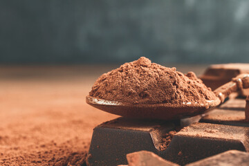 Brown cocoa powder in the spoon and chopped chocolate cubes, on dark background with copy space
