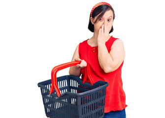 Brunette woman with down syndrome holding supermarket shopping basket covering mouth with hand, shocked and afraid for mistake. surprised expression