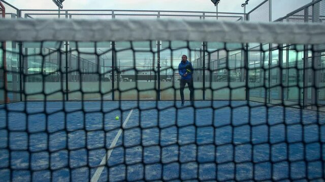 Through grid portrait of paddle tennis man playing on blue court outdoors. One caucasian padle player sportman hitting the ball. Racquet sport game Concept.-Dan