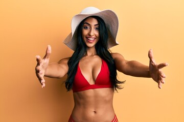 Beautiful hispanic woman wearing bikini and summer hat looking at the camera smiling with open arms for hug. cheerful expression embracing happiness.