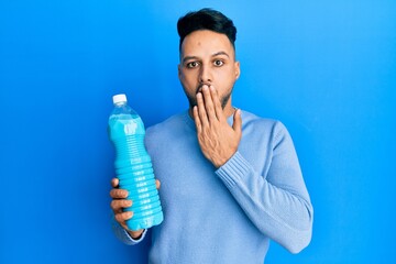 Young arab man holding detergent bottle covering mouth with hand, shocked and afraid for mistake....