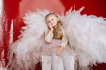 little girl 4 years old in white angel wings on a red latex background in the studio for valentine's day