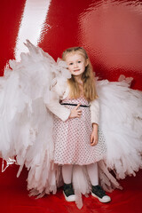 little girl 4 years old in white angel wings on a red latex background in the studio for valentine's day
