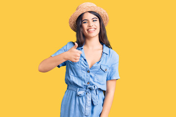 Brunette teenager girl wearing summer hat doing happy thumbs up gesture with hand. approving expression looking at the camera showing success.