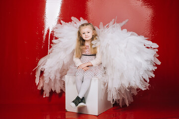 little girl 4 years old in white angel wings on a red latex background in the studio for valentine's day