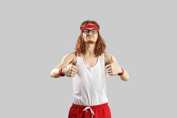 Funny nerdy guy giving you motivation to exercise. Skinny man in gym headband and tank top standing isolated on grey background and showing thumbs up gesture. Sports workout and fitness humour concept - obrazy, fototapety, plakaty