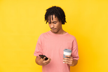 Young african american man isolated on yellow background holding coffee to take away and a mobile