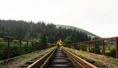 Happy couple of tourists holding hands walking on the railway track on the bridge against the backdrop of a beautiful mountain landscape and smiling. Journey to the mountains.