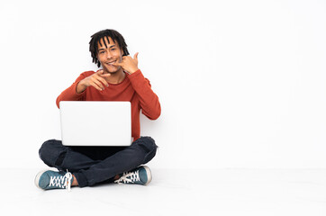 Young african american man sitting on the floor and working with his laptop making phone gesture and pointing front