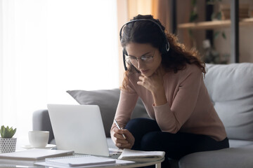 Focused millennial woman in eyeglasses wearing headset with microphone, involved in studying on online lesson using computer, listening educational lecture, writing notes, e-learning concept.