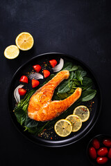 Fried salmon steak with spinach, lemon and cherry tomatoes  served on black plate on wooden table
