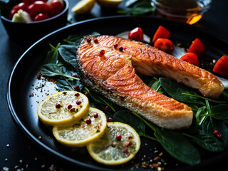 Fried salmon steak with spinach, lemon and cherry tomatoes  served on black plate on wooden table
