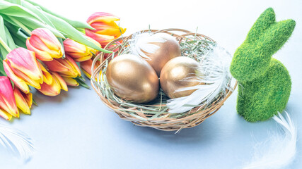 Golden eggs in basket with spring tulips, white feathers on pastel blue background in Happy Easter decoration. Foil minimalist egg design, modern design.