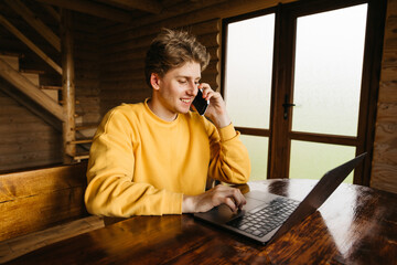 Happy young man working on laptop in the country house in a room with wooden interior and talking on the phone with a smile on his face. Freelance concept.