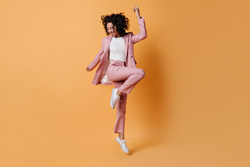 Glad curly girl jumping on yellow background. Full length view of happy lady in pink suit.