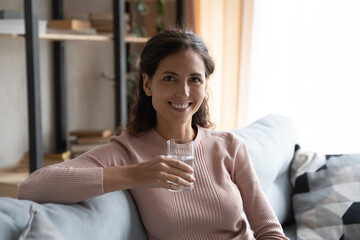 Portrait of happy young woman sitting on sofa, holding glass of fresh mineral water in hands,...