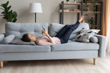 Relaxed millennial woman lying on pillow on cozy couch, involved in communicating distantly with friends in social network or using dating application on smartphone, reading good news at home.