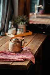 an old hot copper metal teapot sits on a wooden table. Old stylish kitchen in the apartment, sunny daylight from the window. copy space. a red towel.