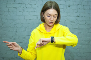 entertained good-looking sociable woman in yellow pointing left with index finger and checking time on watch showing awesome copy space over blue brickwall