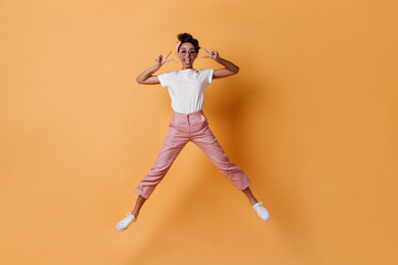 Fototapeta na wymiar Excited girl in pink pants jumping on yellow background. Full length view of mixed race lady showing peace signs.