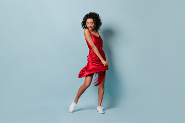 Full length view of playful mixed race girl dancing on blue background. Blissful young woman posing in red dress.
