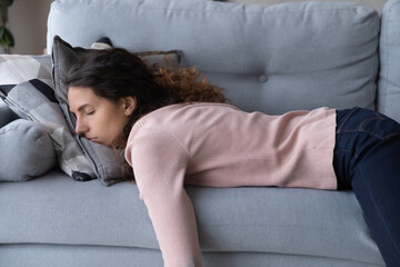 Tired unmotivated young woman falls asleep on cozy couch indoors, having no energy after hard working day. Exhausted caucasian lady napping on comfortable sofa in living room, fatigue concept. - obrazy, fototapety, plakaty