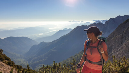 A woman in a hiking outfit with a panoramic view on the haze shrouded valley from the way to Mittagskogel in Austrian Alps. Clear and sunny day. Endless mountain chains. Outdoor activity. Achievement