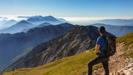 Naklejka premium A man in a hiking outfit with a panoramic view on the haze shrouded valley from the way to Mittagskogel in Austrian Alps. Clear and sunny day. Endless mountain chains. Outdoor activity. Achievement