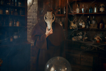 Male exorcist in black hood holds animal skull