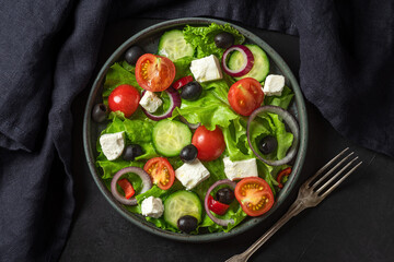 Greek salad with fresh vegetables, feta cheese and olives in a plate with fork and towel on black background. Top view