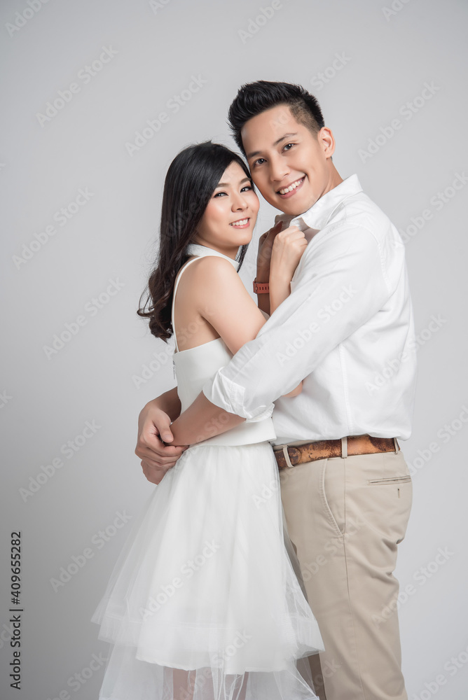 Wall mural happy asian couple in love on gray background. man embracing woman wearing white dress.