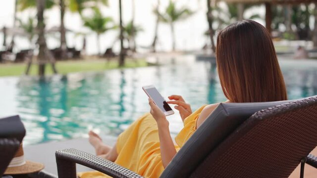 Young woman traveler relaxing and using a mobile phone by a hotel pool while traveling for summer vacation	
