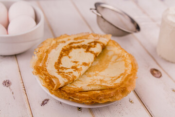 thin delicious round pancakes on a white plate on a wooden table in the kitchen with eggs and flour