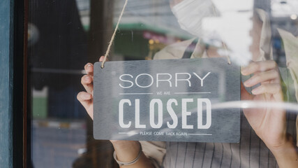 Young Asia girl wear face mask turning a sign from open to closed sign on glass door cafe after coronavirus lockdown quarantine. Owner small business, food and drink, business financial crisis concept