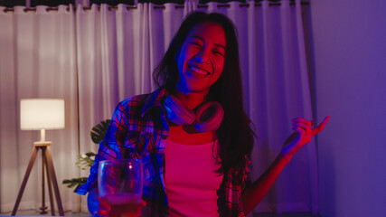 Young Asia lady drinking beer having fun happy moment disco neon night party event online celebration via video call in living room at home. Social distancing, quarantine for coronavirus prevention.
