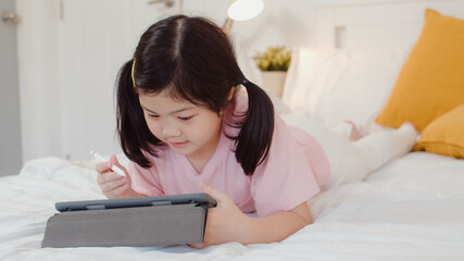Young Asian girl drawing at home. Asia japanese woman child kid relax rest fun happy using tablet draw cartoon before sleep lying on bed, feel comfort and calm in bedroom at night concept.