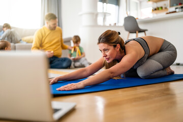 Young woman, mother exercising at home in living room, father playing with kids in background.